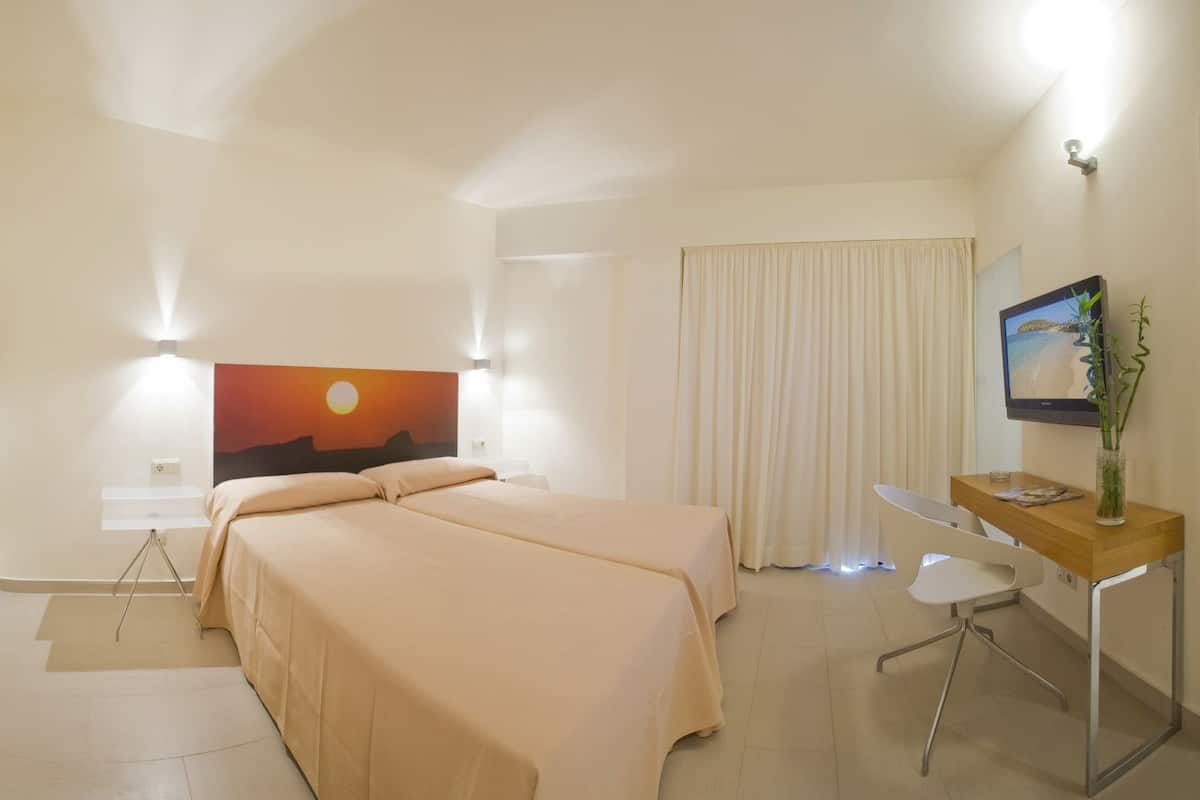 A hotel room with two beds, a desk, a TV, and a painting of a sunset.