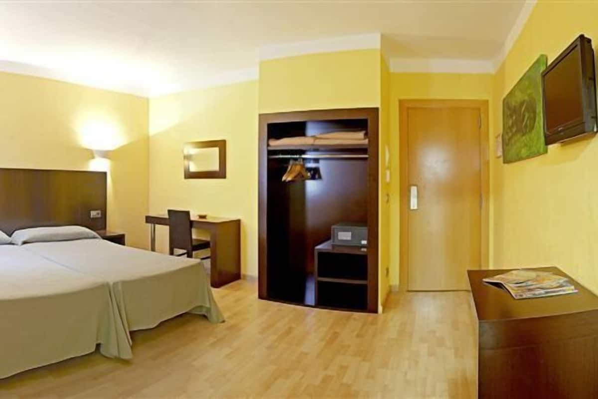 A hotel room with a double bed, a desk with a chair, a wardrobe, and a TV on the wall.