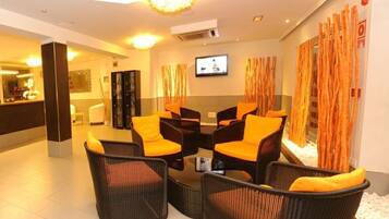 A modern hotel lobby with a reception desk, seating area with orange chairs, a television, and a decorative plant.