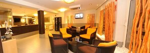 A modern hotel lobby with a reception desk, seating area with orange chairs, a television, and a decorative plant. - Hotel Florencio (Sant Antoni de Portmany)