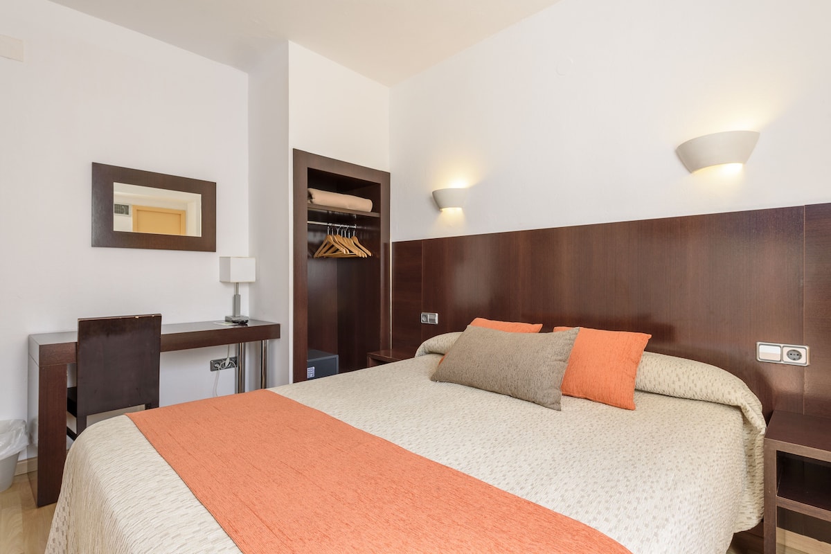 A hotel room with a bed, a desk, a chair, a wardrobe, and a mirror.