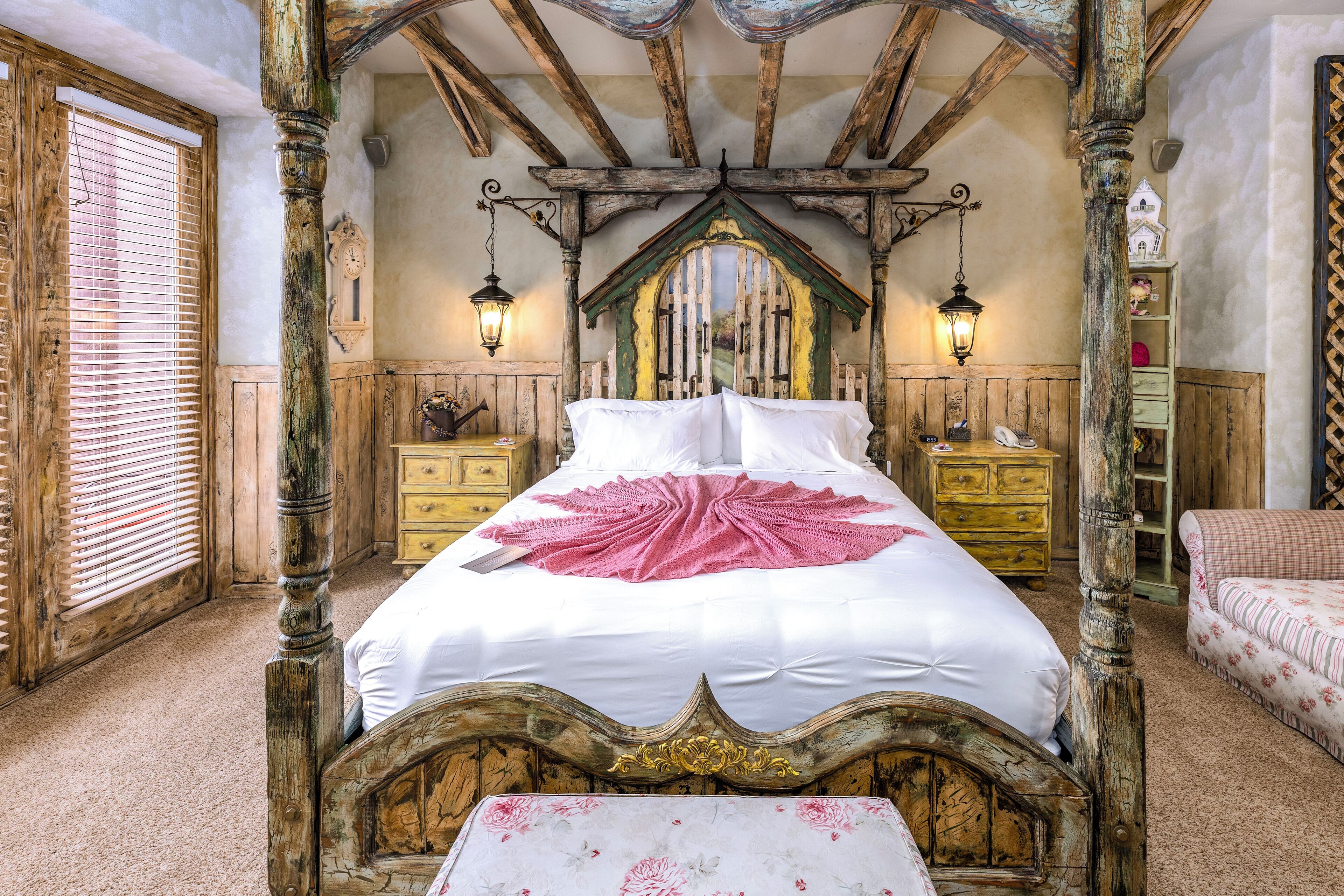 A traditional bedroom with a four-poster bed, wooden headboard, and a canopy. There are two bedside tables with lamps, a wooden dresser, and a small table with a floral tablecloth.