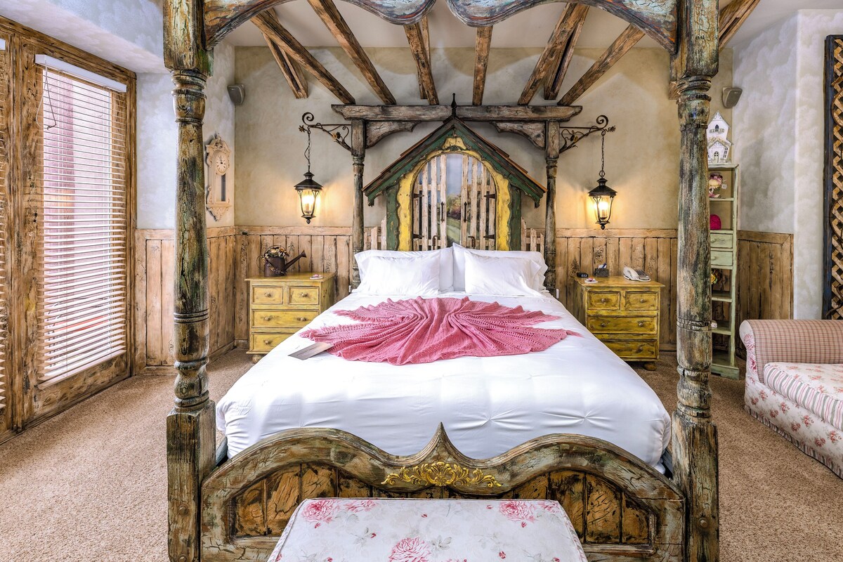 Un dormitorio tradicional con cama con dosel, cabecera de madera y dosel. Hay dos mesitas de noche con lámparas, un aparador de madera y una pequeña mesa con mantel floral.