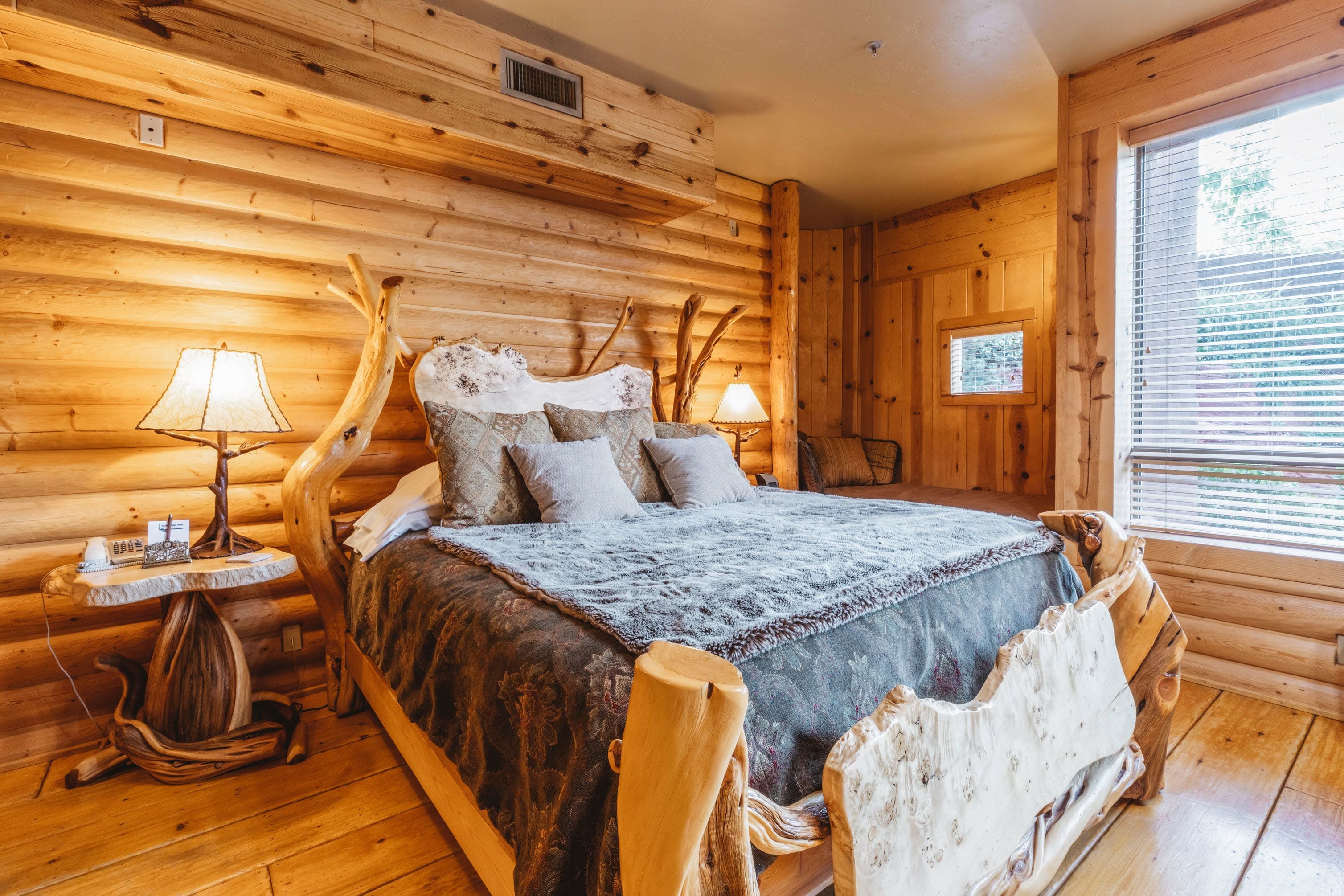 Une chambre à coucher dans un chalet en rondins, avec un lit, une tête de lit en bois, deux tables de chevet avec des lampes et une fenêtre avec des stores.