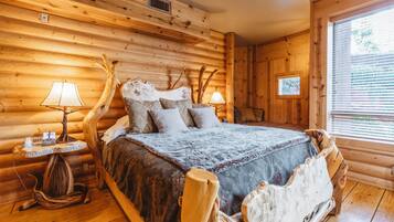 Une chambre à coucher dans un chalet en rondins, avec un lit, une tête de lit en bois, deux tables de chevet avec des lampes et une fenêtre avec des stores.