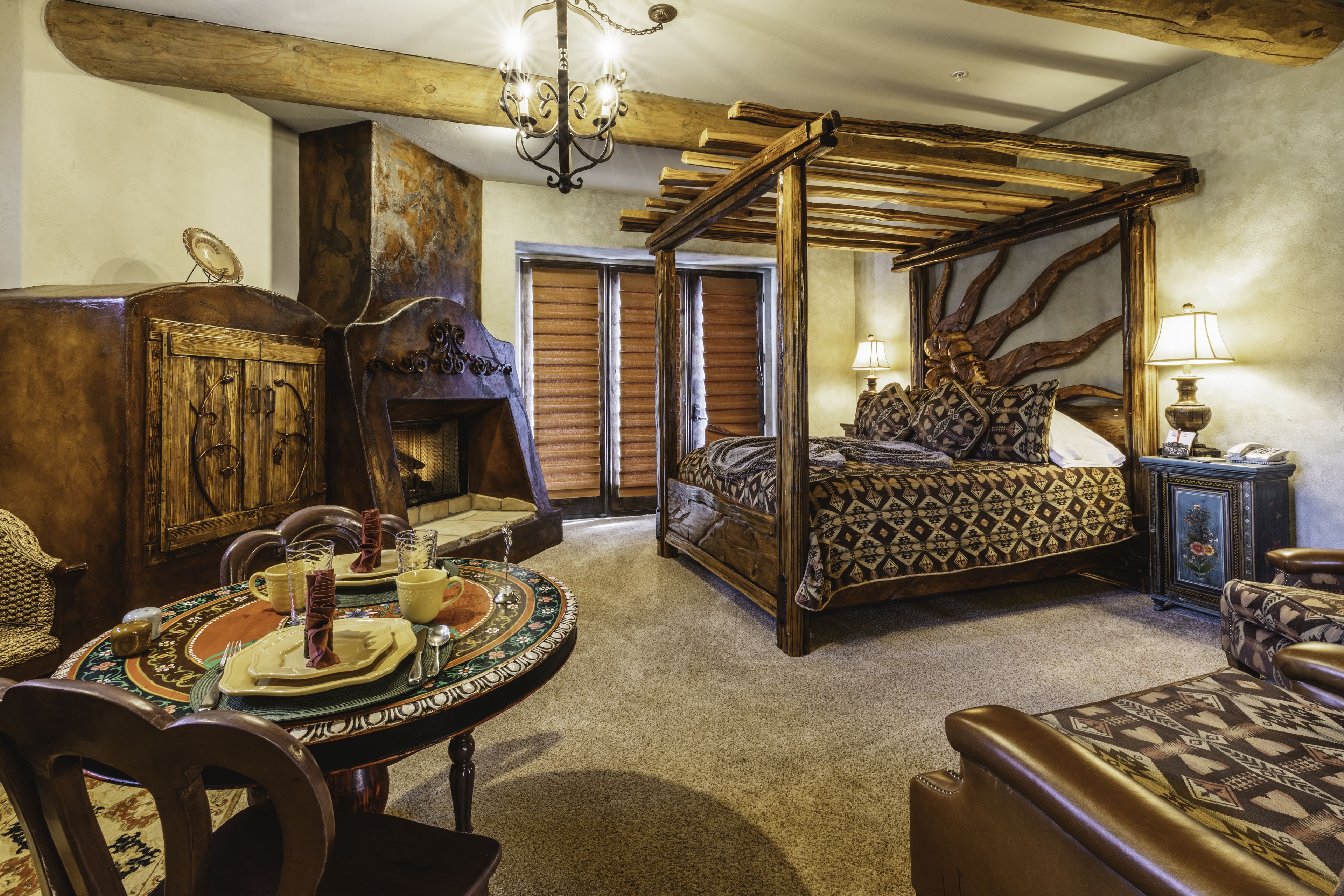 A bedroom with a four-poster bed, a wooden wardrobe, a round dining table with a decorative centerpiece, and a fireplace.