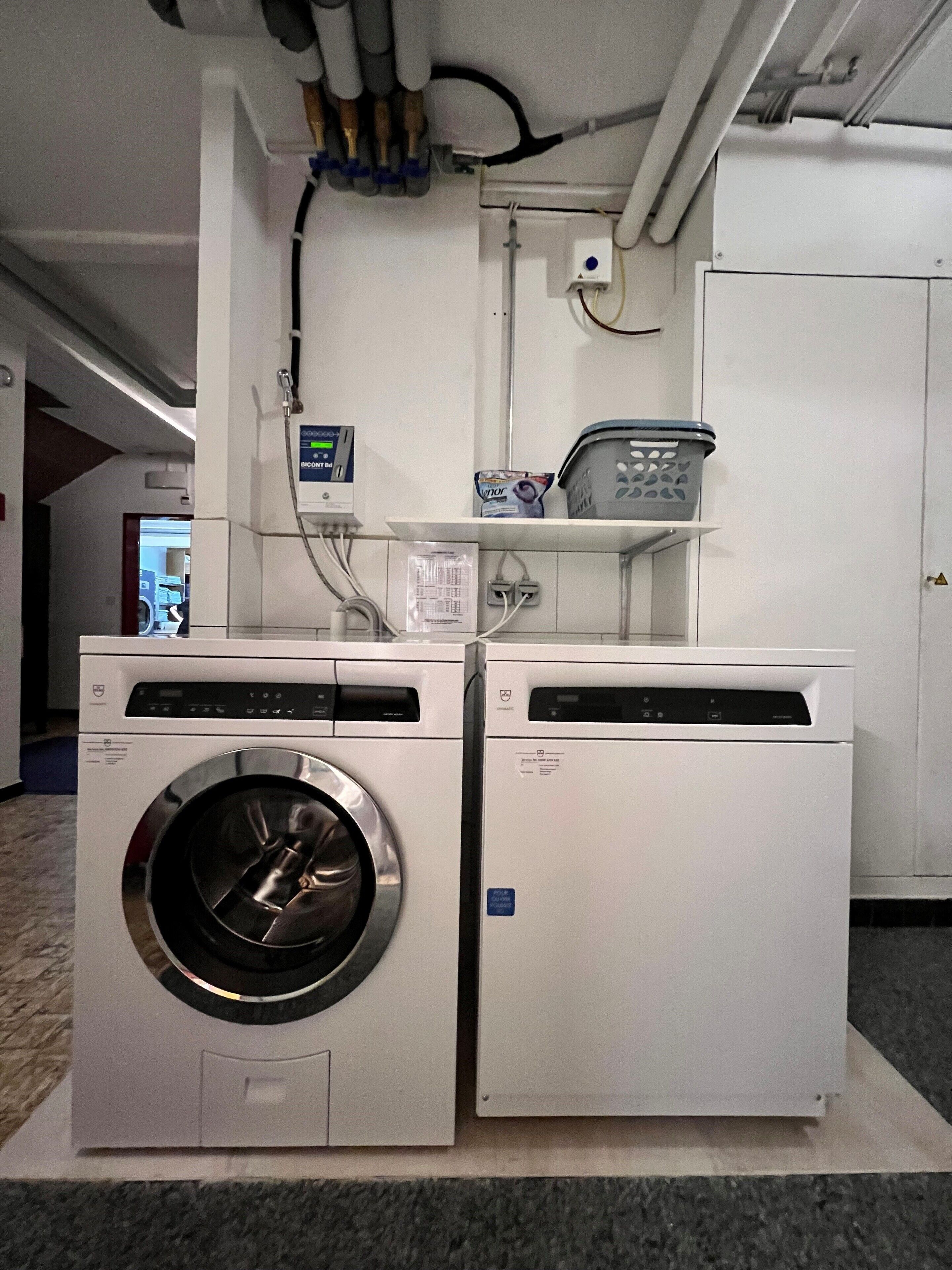 A laundry area with two white front-loading washing machines, a control panel, and a shelf with laundry baskets.
