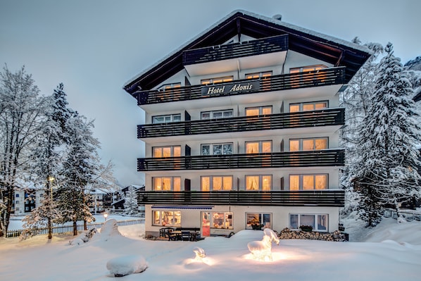 A multi-story hotel with balconies, a sign reading "Hotel Adonis," and snow-covered surroundings. - Adonis (Zermatt)