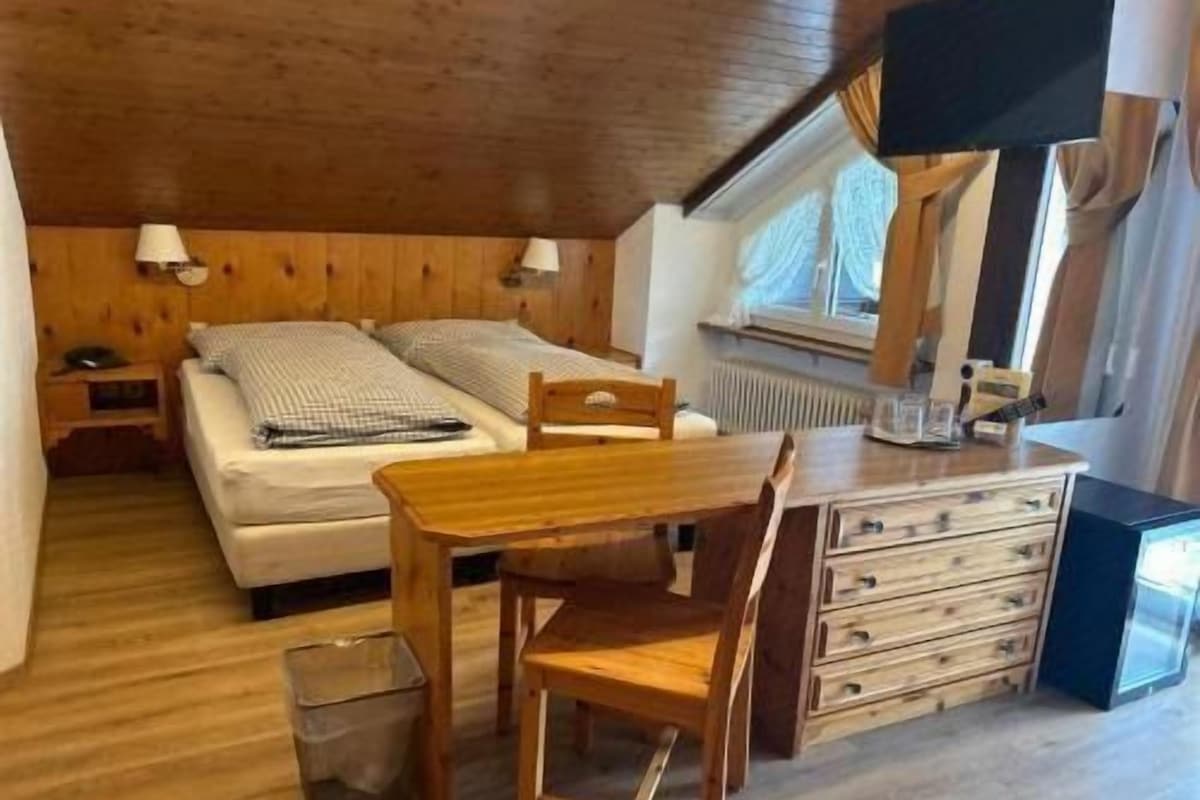 Une petite pièce confortable, avec un lit, une table en bois, une chaise, une commode et une fenêtre.