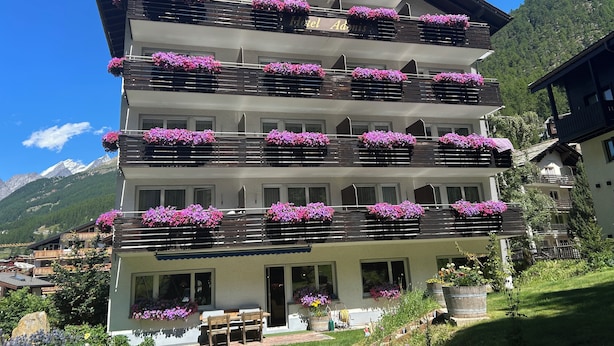 A multi-story hotel with balconies adorned with purple flowers, a mountain backdrop, and a well-maintained garden area.