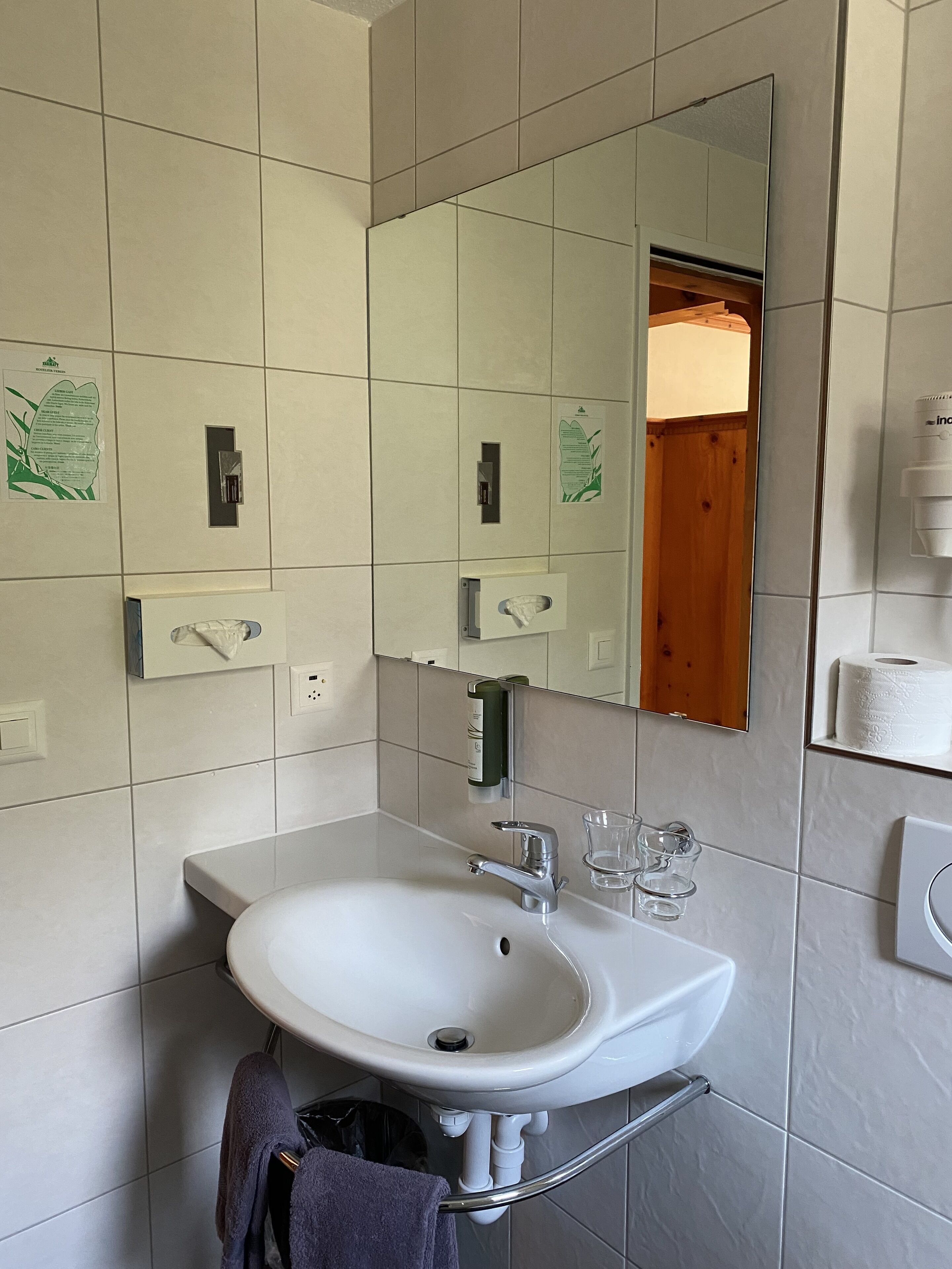 A bathroom with a white sink, mirror, and tiled walls. There are paper towel dispensers, a soap dispenser, and a glass of water on the counter.