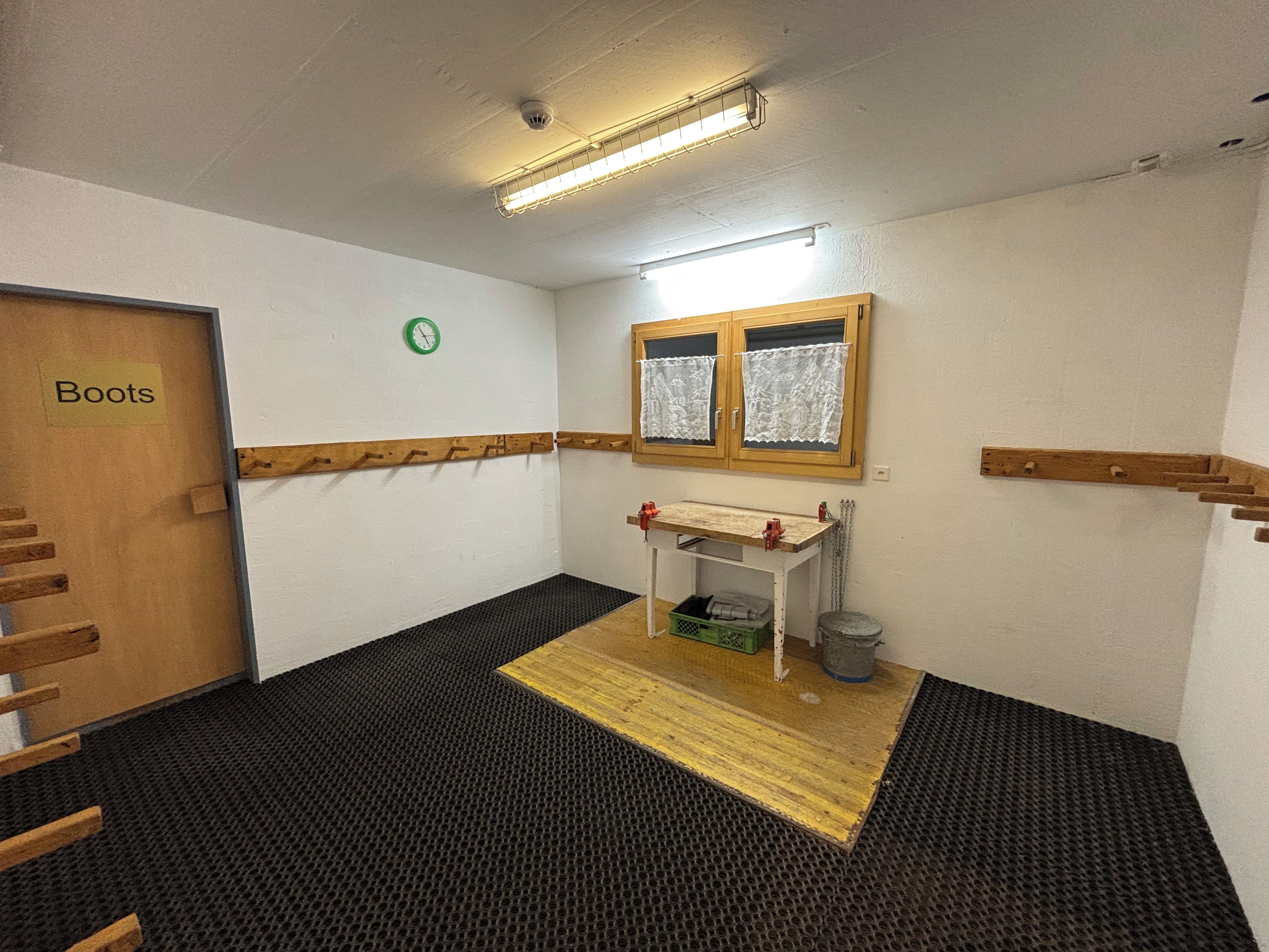 A small room with a wooden door labeled 'Boots,' a small table with a green basket, a window with lace curtains, and a wall-mounted clock.