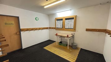 A small room with a wooden door labeled 'Boots,' a small table with a green basket, a window with lace curtains, and a wall-mounted clock.