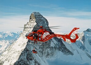 A red helicopter with stars and the letters "HB-ZEF" flying over a snow-covered mountain. - Adonis (Zermatt)