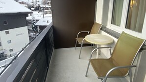 A balcony with two chairs and a small table, overlooking a snowy mountain and buildings.