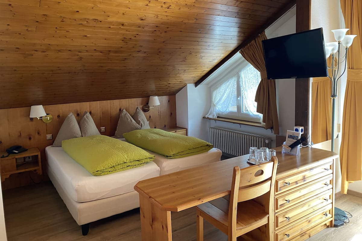Ein gemütliches Schlafzimmer mit Holzbalkendecke, einem Bett mit grün-beigen Kissen, einem Holztisch und einem an der Wand montierten Fernseher.