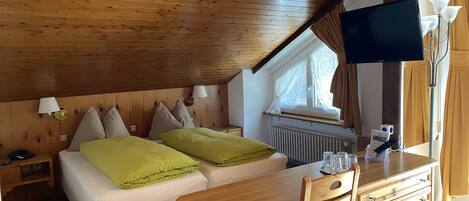 A cozy bedroom with a wooden ceiling, a bed with green and beige pillows, a wooden desk, and a television mounted on the wall.