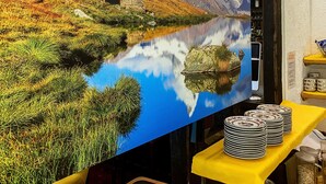 A room with a large wall mural of a mountain landscape and a lake, a yellow table with stacked plates, and a tray with utensils.