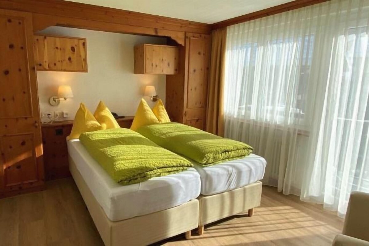A double bed with pillows, a nightstand, a lamp, and a window with sheer curtains.