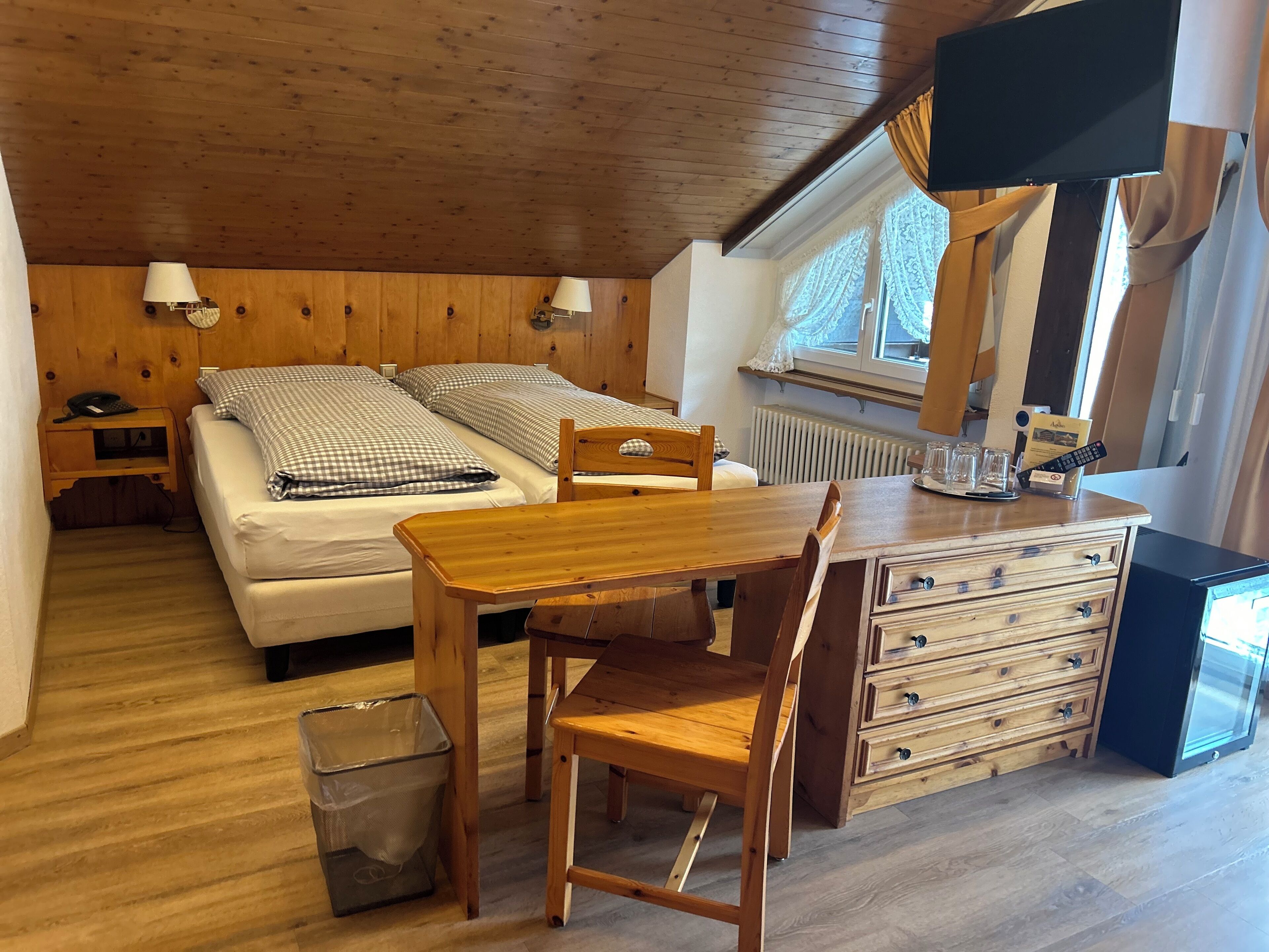 Una habitación acogedora con dos camas, una mesa de madera, una silla, un aparador, un cesto de basura, un calentador y un televisor.