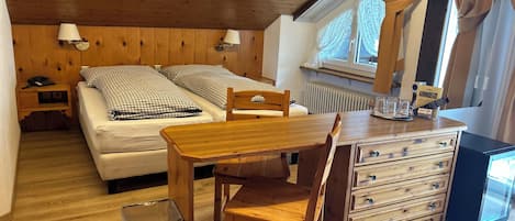 Una habitación acogedora con dos camas, una mesa de madera, una silla, un aparador, un cesto de basura, un calentador y un televisor.