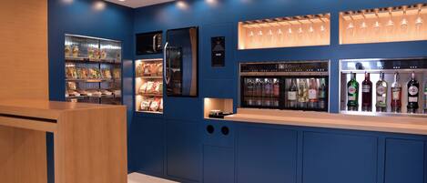 A modern bar area with a blue color scheme, featuring a refrigerator, shelves with snacks, and a display of bottles.