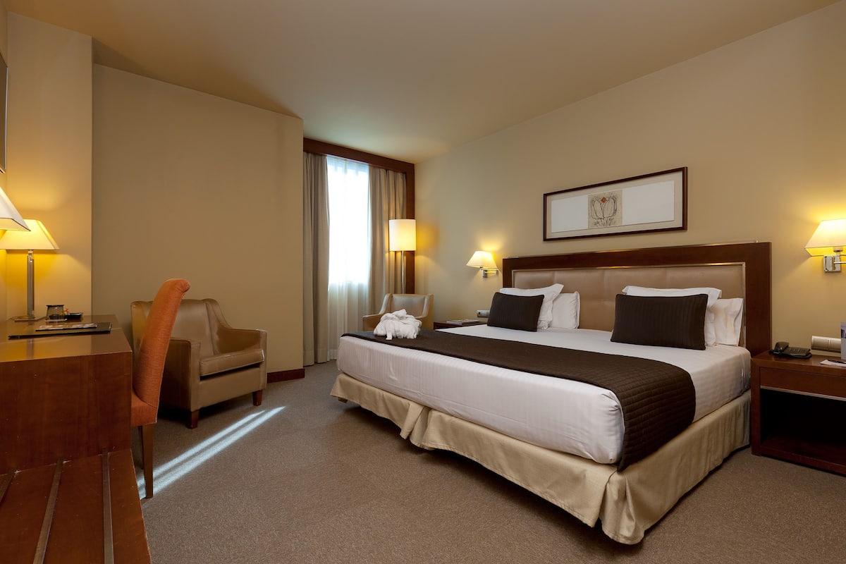 Une chambre d’hôtel avec un grand lit, deux lampes de chevet, un bureau avec une lampe, une chaise et une fenêtre avec des rideaux.