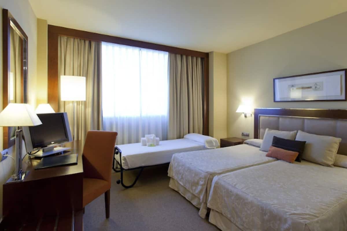 Une chambre d’hôtel avec deux lits, un bureau avec un ordinateur, une chaise et une fenêtre avec des rideaux.