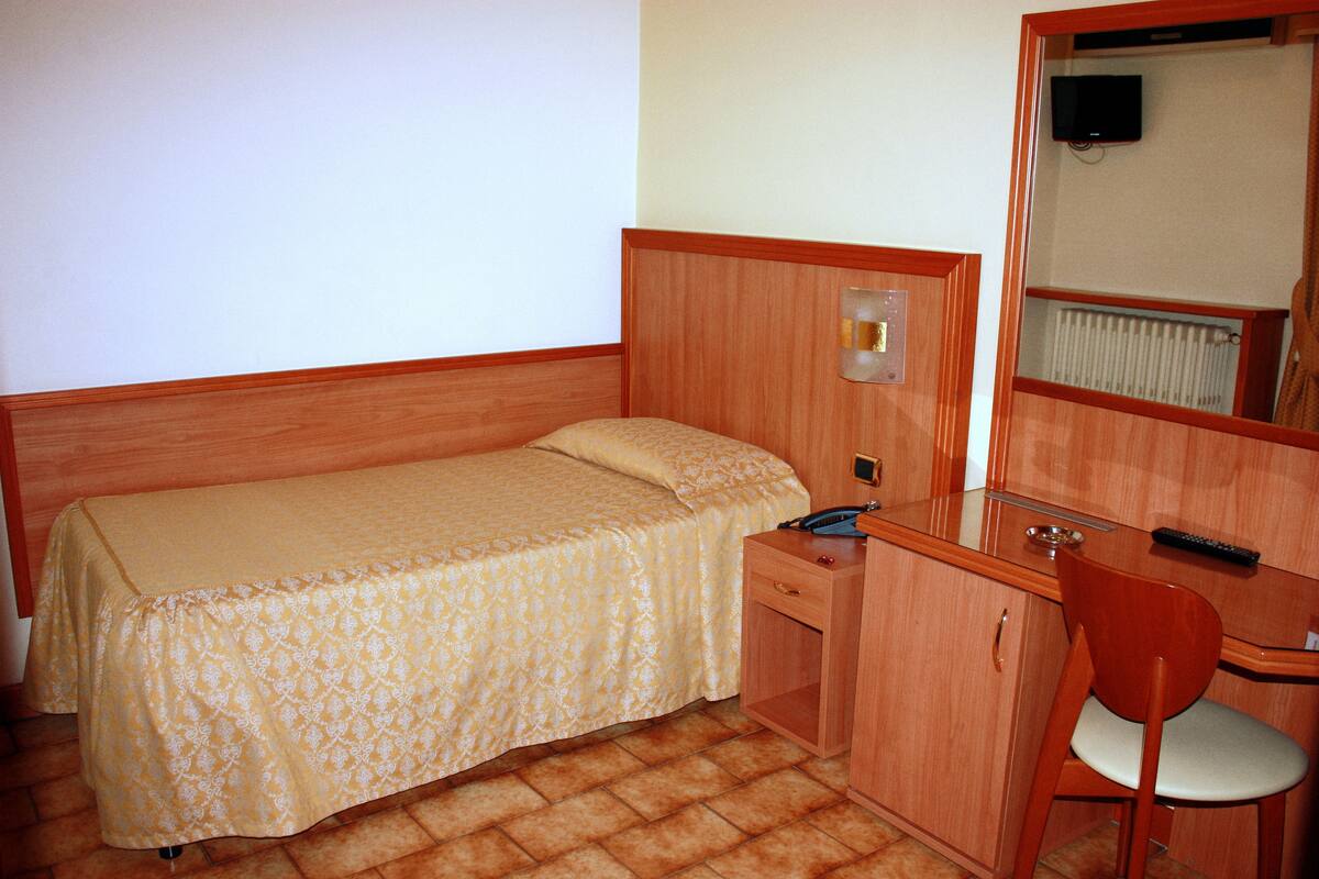 Une petite chambre à coucher, bien rangée, avec un lit simple, un bureau en bois, une chaise, un miroir et un téléviseur.