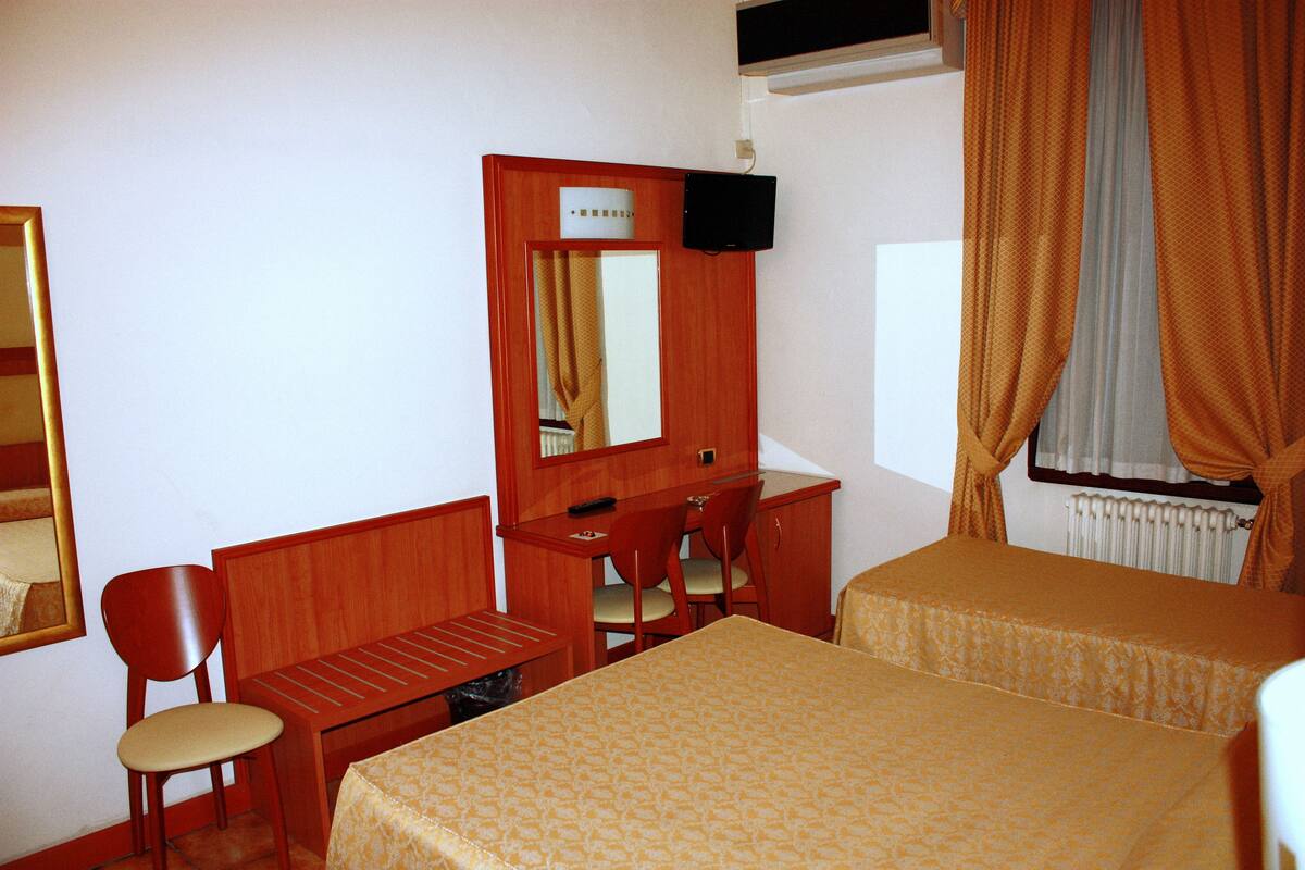 Une chambre d’hôtel avec un bureau en bois, deux lits simples, une chaise, un miroir et une fenêtre avec des rideaux.