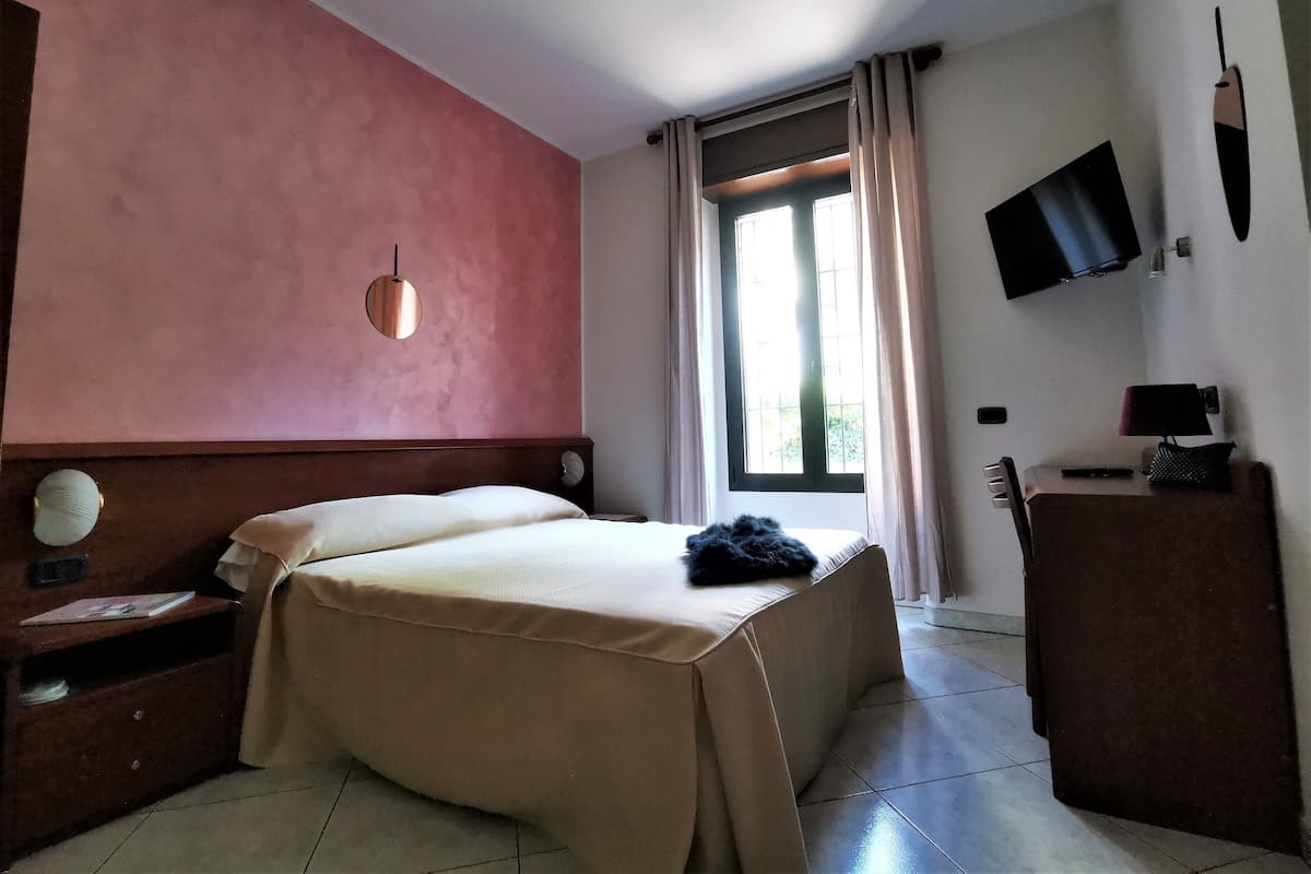 A hotel room with a bed, bedside table, lamp, TV, and a window with curtains.