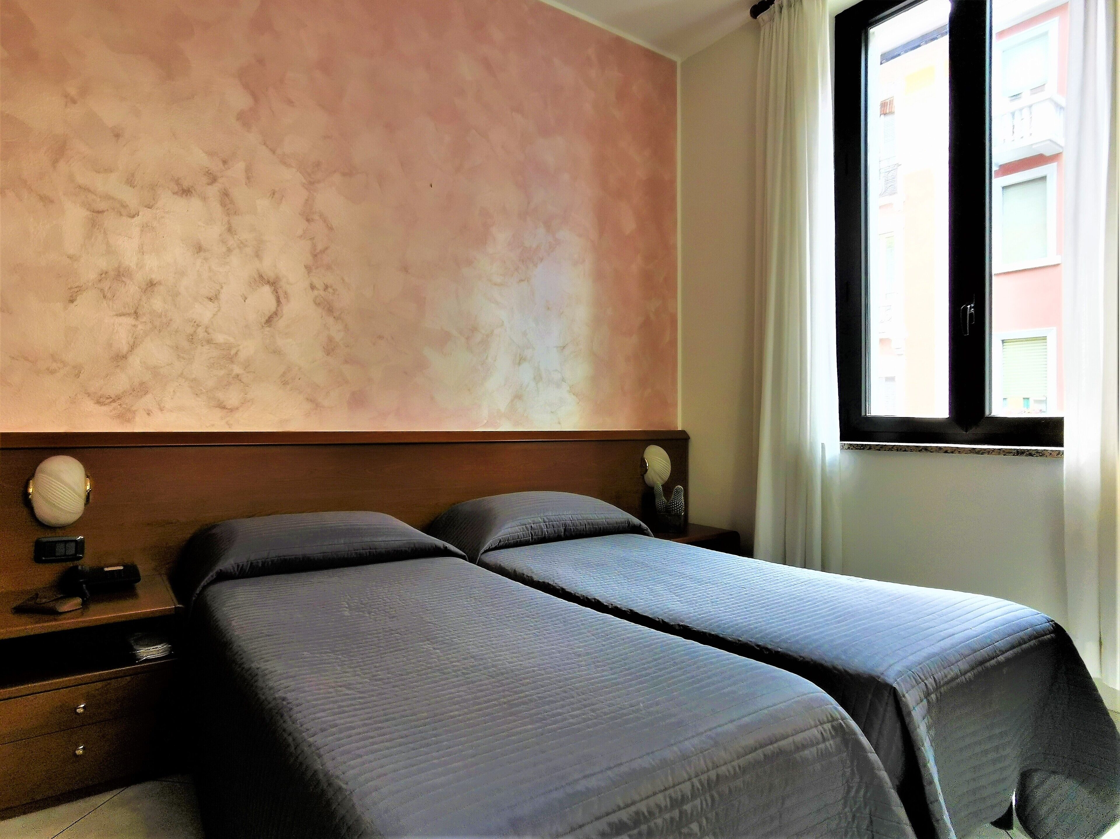 Ein Zimmer mit zwei Betten, einem hölzernen Kopfteil, einem Nachttisch mit Lampe und einem Fenster mit Vorhängen.