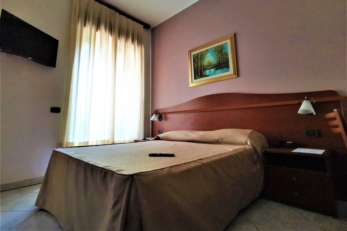 A hotel room with a bed, a nightstand, a lamp, a painting on the wall, and a window with curtains.