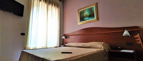 A hotel room with a bed, a nightstand, a lamp, a painting on the wall, and a window with curtains.