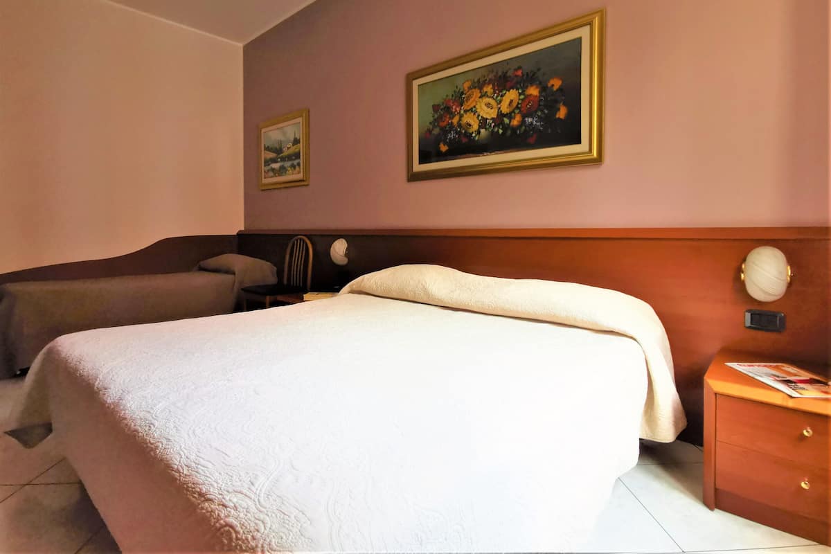 A hotel room with a large bed, a smaller bed, a nightstand with a lamp, a chair, and two framed paintings on the wall.