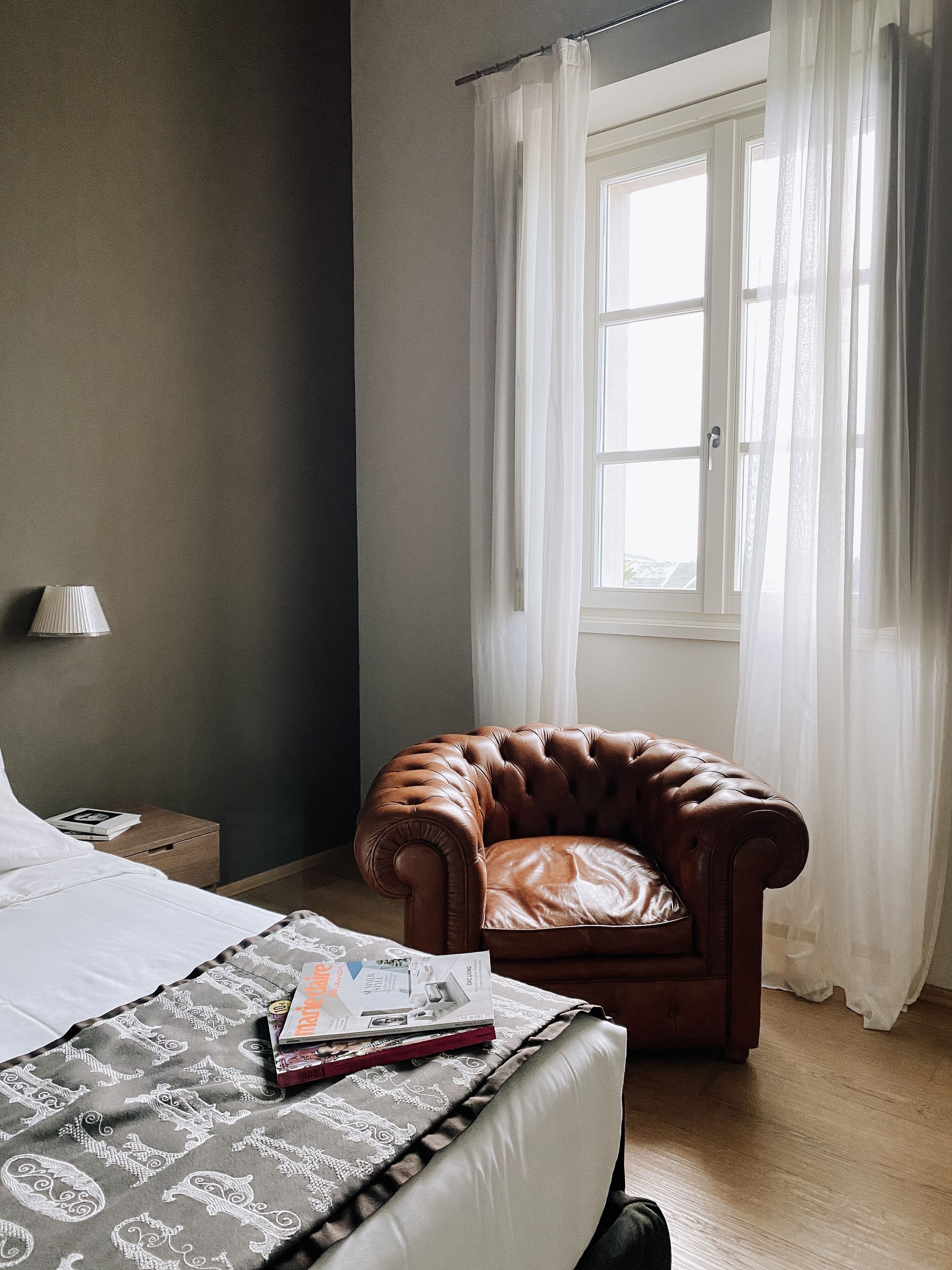 A bedroom with a bed, a brown leather armchair, a window with sheer curtains, and a bedside table with a lamp and a book.