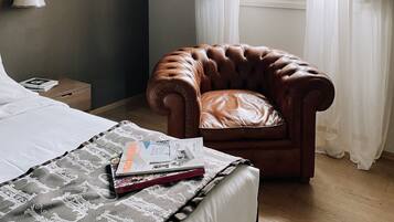 A bedroom with a bed, a brown leather armchair, a window with sheer curtains, and a bedside table with a lamp and a book.