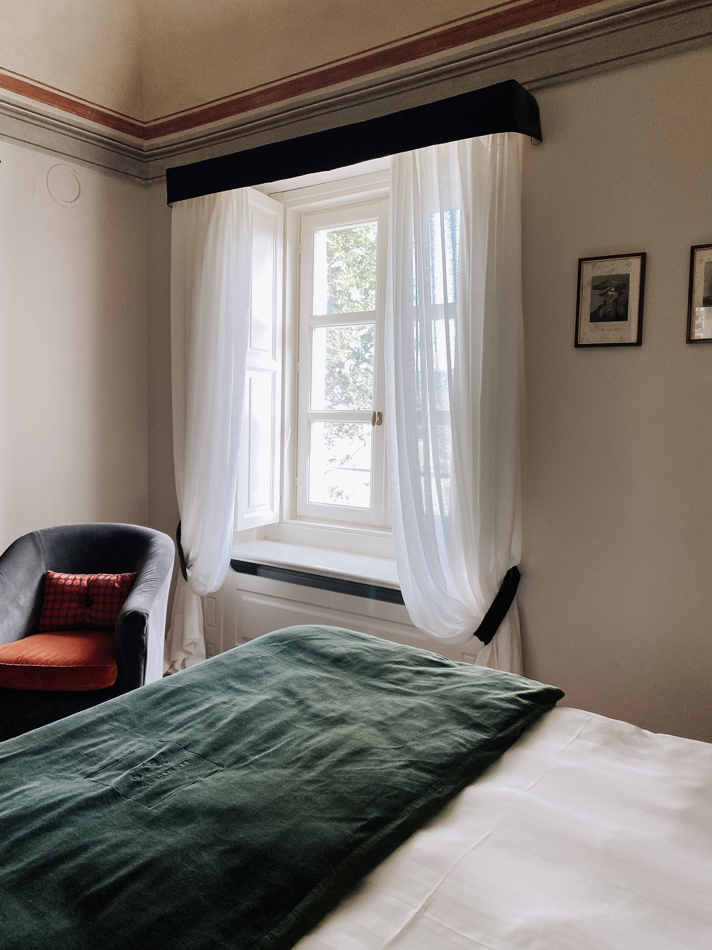 Una habitación con una cama, una silla, un ventanal con cortinas traslúcidas y un cuadro en la pared.