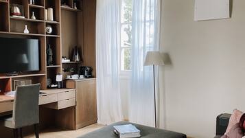 Una sala de estar moderna con un sofá, un reposapiés y un escritorio con computadora. Hay un televisor, una estantería y una ventana con cortinas.