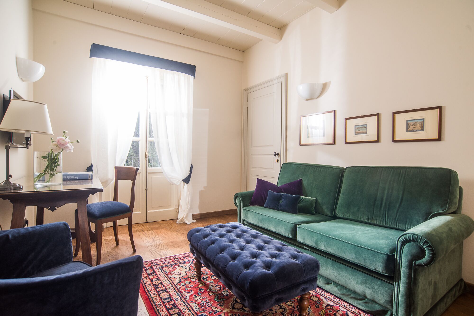 Un salón con un sofá verde, dos sillones, un reposapiés azul, una mesa de centro de madera y una alfombra estampada.