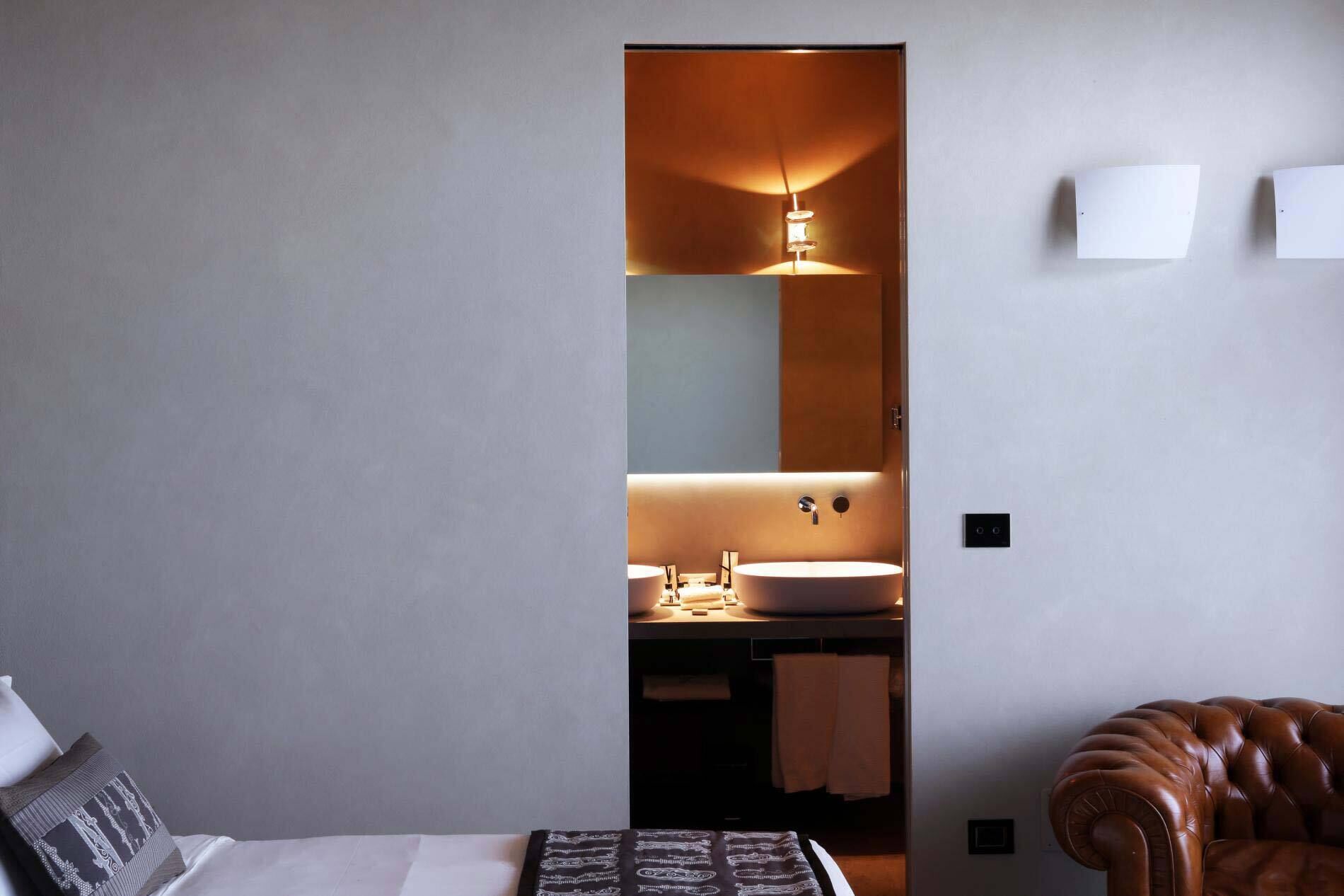 Una habitación de hotel moderna con una cama grande, un sillón de madera y un baño con un espejo amplio y dos lavamanos.