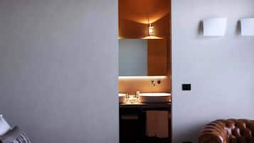 Una habitación de hotel moderna con una cama grande, un sillón de madera y un baño con un espejo amplio y dos lavamanos.