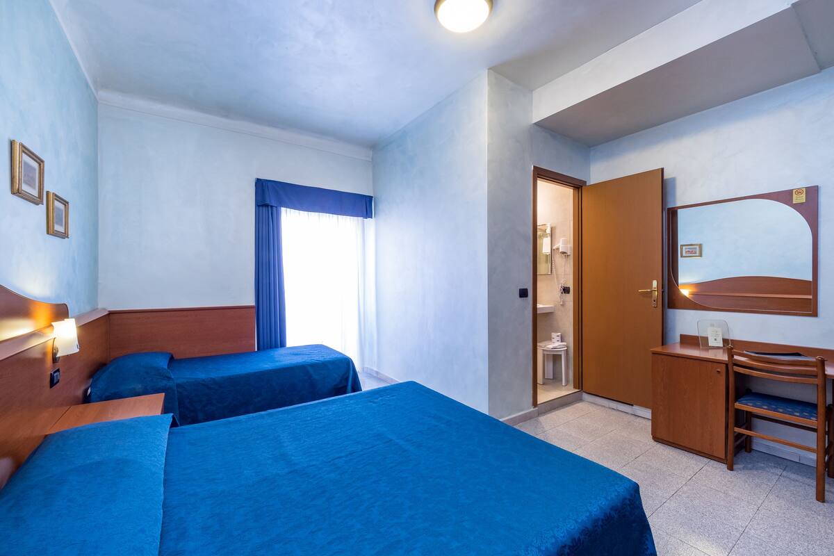 A hotel room with two beds, a desk, a mirror, and a bathroom visible through an open door.