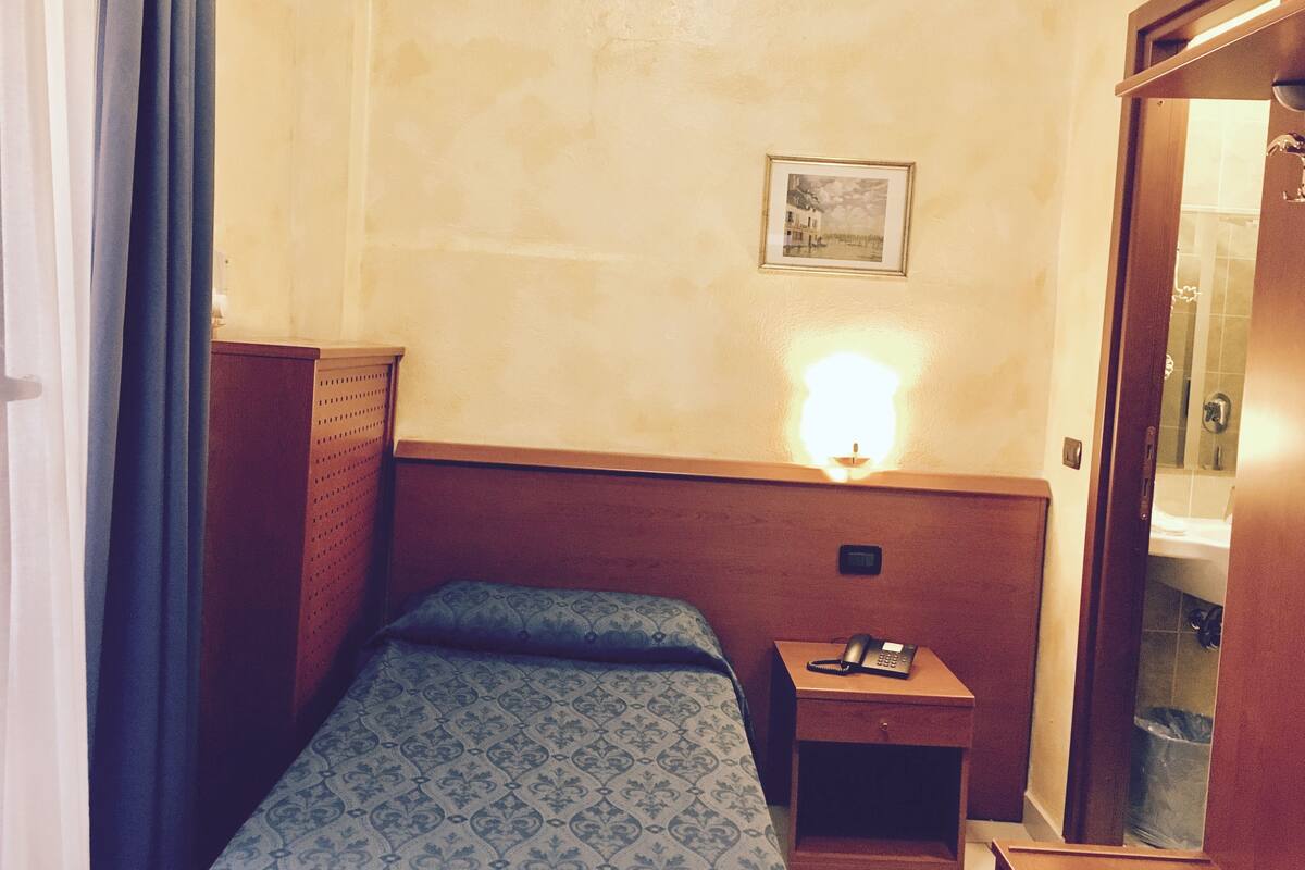A small hotel room with a single bed, a nightstand with a phone, a wall-mounted light, and a framed picture.