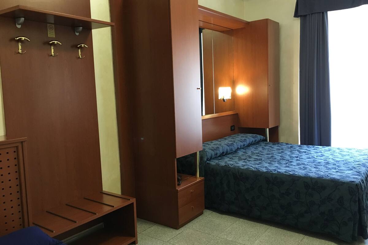 A compact hotel room with a bed, a wardrobe, a mirror, and a small seating area.