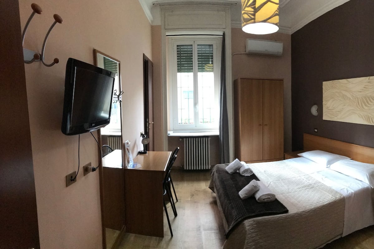 A hotel room with a bed, bedside table, TV, and a window with blinds.