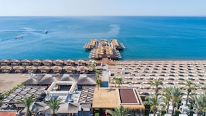 An aerial view of a beach resort with multiple thatched-roof structures, sun loungers, and a pier extending into the sea. - Titanic Deluxe Lara - All inclusive (Antalya)