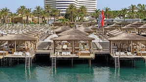 A resort with multiple overwater cabanas, a pool, and a large hotel building in the background. - Titanic Deluxe Lara - All inclusive (Antalya)