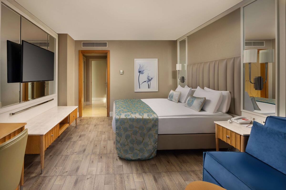 Quarto de hotel com cama, mesa de cabeceira, televisão e escrivaninha com cadeira.