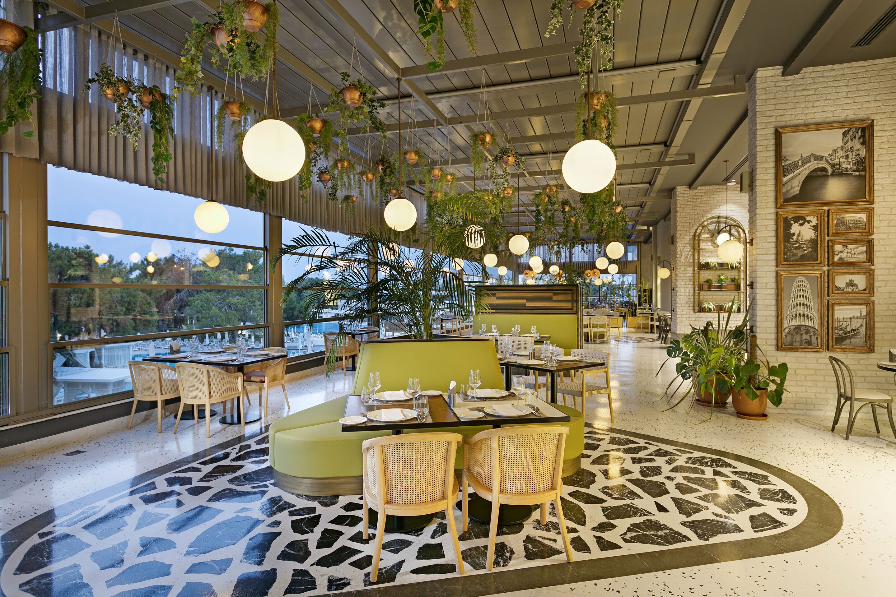A modern restaurant with round tables, green seating, hanging plants, and large windows offering a view of the outdoors.